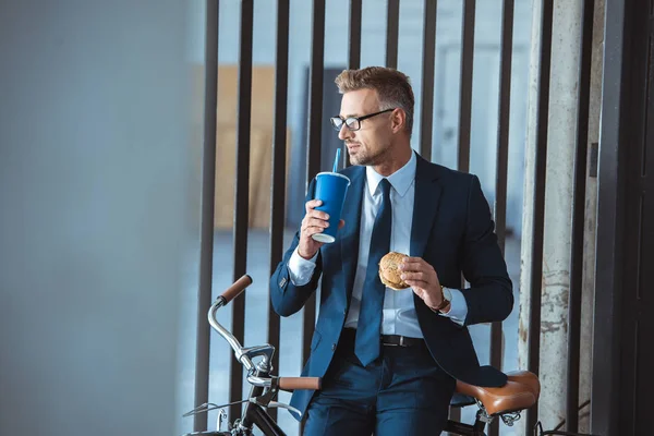 Homme d'affaires tenant hamburger et boire de tasse en papier tout en étant assis sur le vélo et regardant loin — Photo de stock