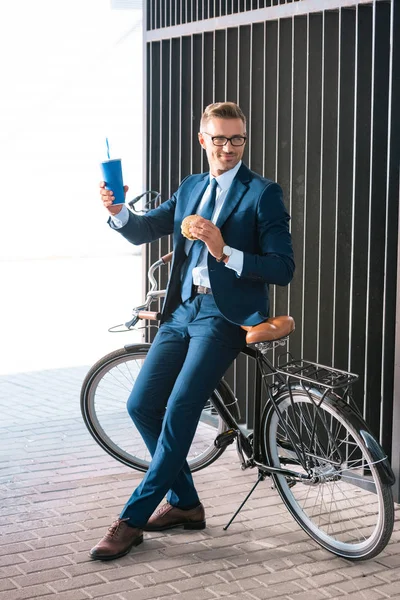 Guapo sonriente hombre de negocios sosteniendo taza desechable y hamburguesa mientras está sentado en bicicleta — Stock Photo