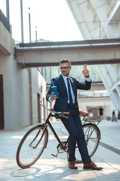 Улыбающийся бизнесмен, сидящий на велосипеде, держащий бумажную чашку и показывающий два пальца на улице — стоковое фото