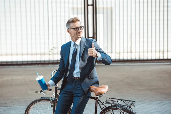 Улыбающийся бизнесмен, сидящий на велосипеде, держащий бумажную чашку и показывающий большой палец на улице — стоковое фото