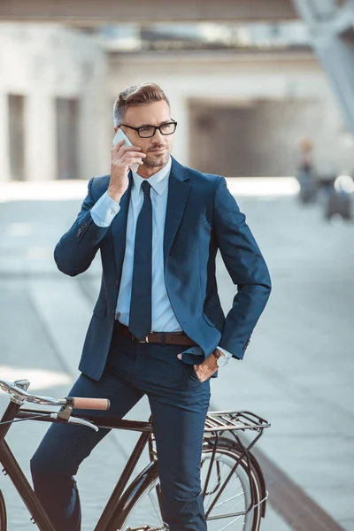 Handsome middle aged businessman in formal wear and eyeglasses sitting on bicycle and talking by smartphone — Stock Photo