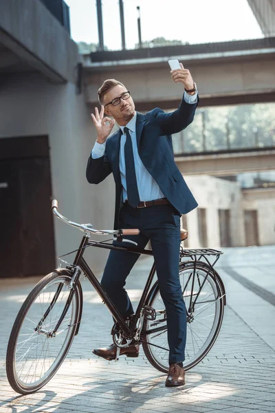 Усміхнений бізнесмен, показуючи знак ОК на смартфон, сидячи на велосипеді на вулиці — стокове фото