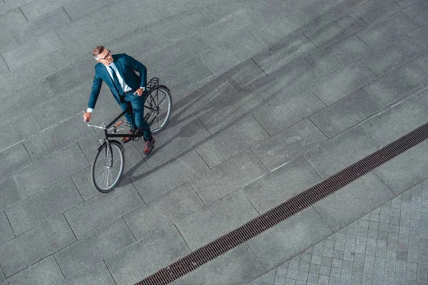 Високий кут зору красивого бізнесмена в костюмі і окулярах, сидячи на велосипеді і дивлячись в сторону — стокове фото