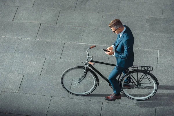 Високий кут зору бізнесмена в формальному носінні за допомогою смартфона, стоячи з велосипедом на вулиці — стокове фото
