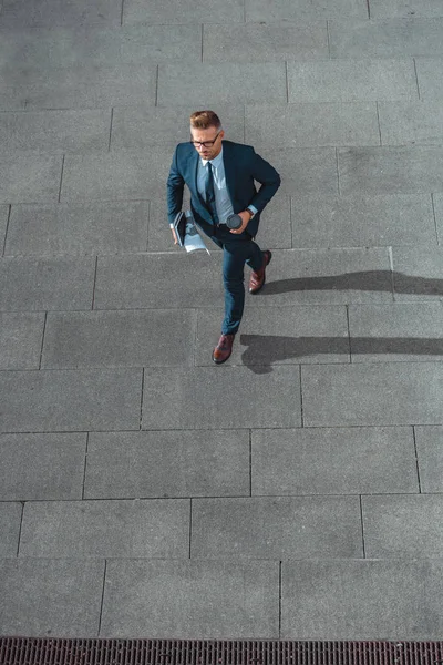 Vista ad alto angolo di uomo d'affari con tazza di carta e giornale che cammina sulla strada — Foto stock