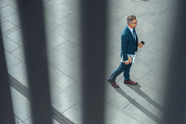 Vista de ángulo alto de hombre de negocios con café para ir y periódico caminando por la calle - foto de stock