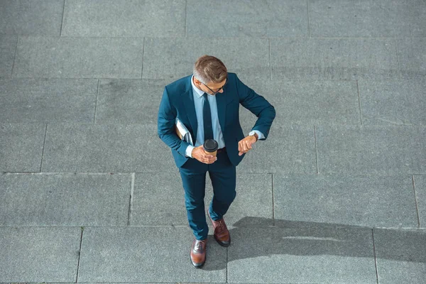 Successful middle aged businessman holding coffee to go and checking wristwatch on street — Stock Photo