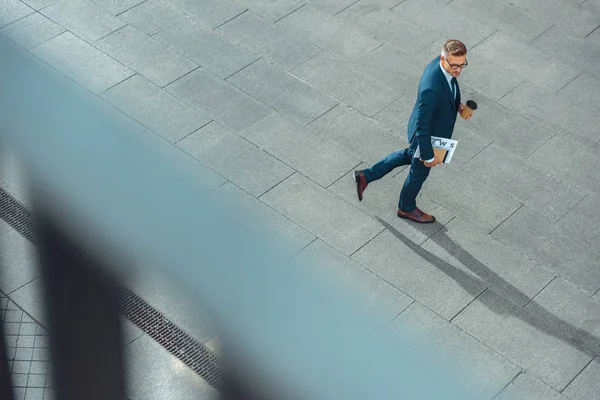 Vista de ángulo alto de hombre de negocios con taza de papel y periódico caminando por la calle - foto de stock