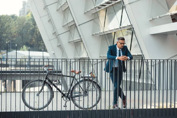 Exitoso hombre de negocios de mediana edad en ropa formal apoyado en barandilla cerca de la bicicleta - foto de stock