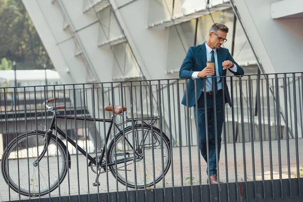 Exitoso hombre de negocios de mediana edad en uso formal sosteniendo la taza de papel y comprobando el reloj de pulsera mientras está de pie cerca de la bicicleta - foto de stock