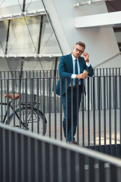 Businessman in formal wear holding coffee to go and adjusting eyeglasses while standing near bike on street — Stock Photo