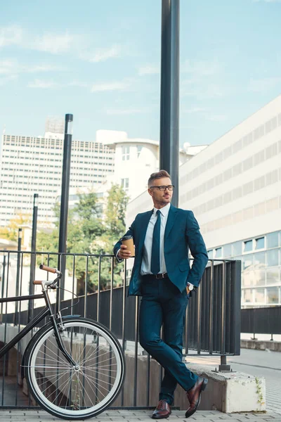 Guapo hombre de negocios sosteniendo café para ir y mirar hacia otro lado mientras está parado cerca de la bicicleta en la calle — Stock Photo