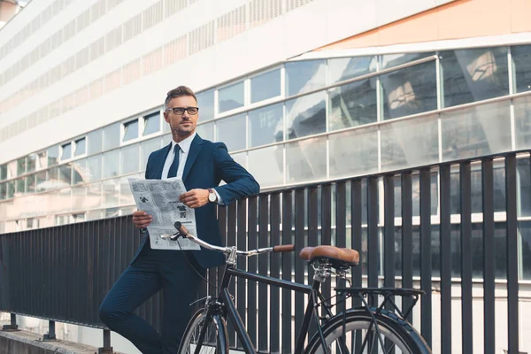 Erfolgreicher Geschäftsmann in offizieller Kleidung hält Zeitung in der Hand und schaut weg, während er mit dem Fahrrad auf der Straße steht — Stockfoto