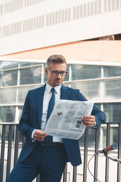 Schöner Geschäftsmann mittleren Alters in Anzug und Brille, der auf der Straße Zeitung liest — Stockfoto