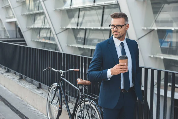 Hombre de negocios guapo en traje y gafas con taza de papel y mirando hacia otro lado mientras está de pie con la bicicleta en la calle - foto de stock