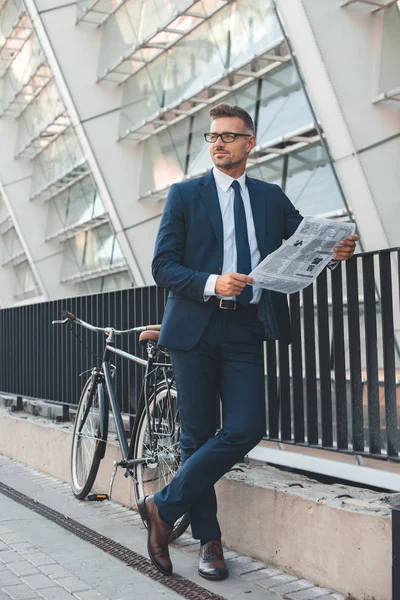 Handsome buisnessman in eyeglasses holding newspaper and looking away while standing with bicycle on street — Stock Photo