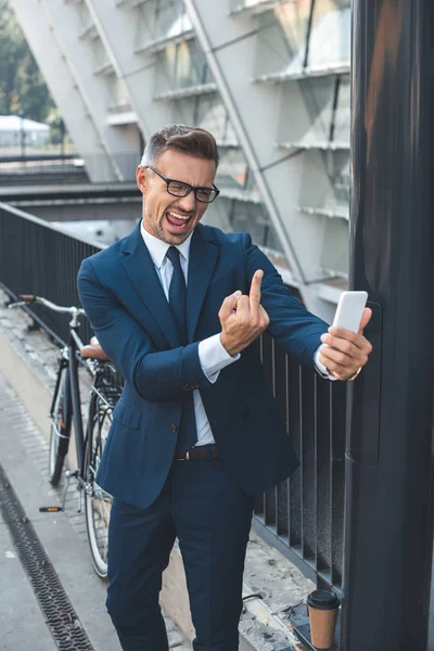 Emotional middle aged businessman in formal wear and eyeglasses holding smartphone and showing middle finger — Stock Photo