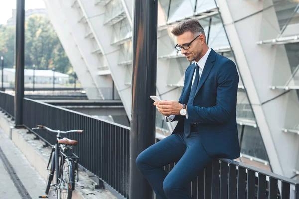 Smiling middle aged businessman in eyeglasses using smartphone while sitting on street — Stock Photo