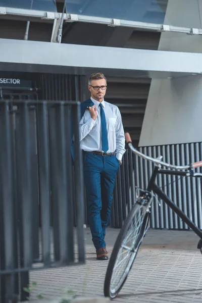 Handsome businessman in eyeglasses holding suit jacket and going to bicycle parked on street — Stock Photo