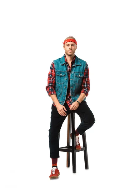 Модный хипстер в джинсовой жилетке и красной клетчатой рубашке сидит на стуле изолирован на белом — стоковое фото