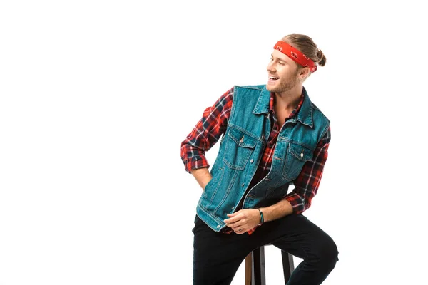 Happy hipster man in denim vest and red checkered shirt looking away on chair isolated on white — Stock Photo