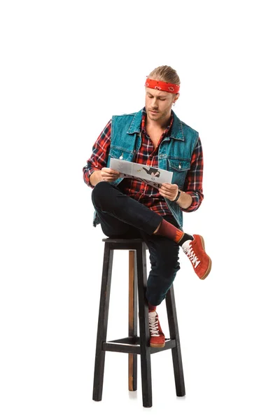 Stylish hipster man in denim vest reading travel newspaper on chair isolated on white — Stock Photo