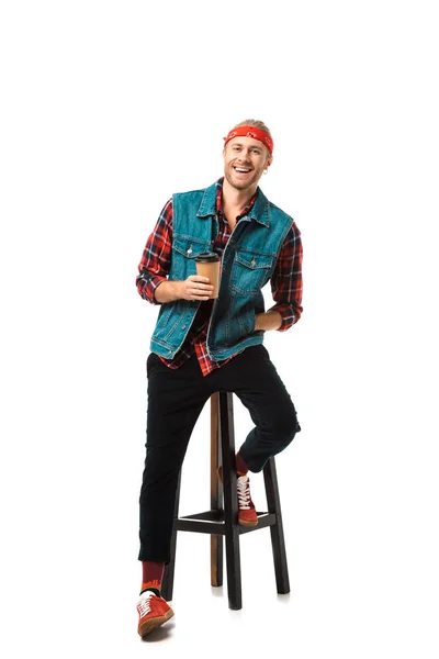 Homem alegre na cabeça e colete jeans sentado na cadeira com xícara de café isolado no branco — Fotografia de Stock