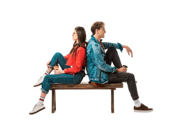 Vista lateral do casal hipster com xícaras de papel de café sentado de costas para trás no banco isolado em branco — Fotografia de Stock