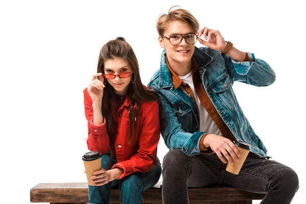 Happy hipster couple with coffee cups adjusting eyeglasses and sitting on bench isolated on white — Stock Photo