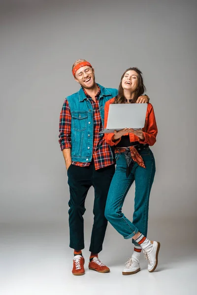 Riéndose pareja hipster de pie con el ordenador portátil en gris - foto de stock