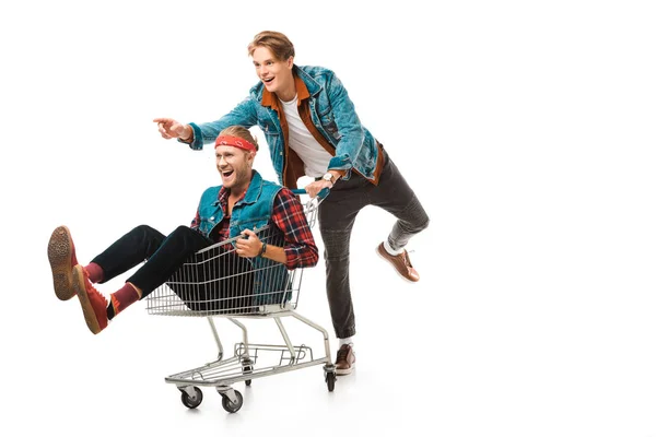 Cheerful hipster pointing by finger and carrying shopping trolley with male friend isolated on white — Stock Photo