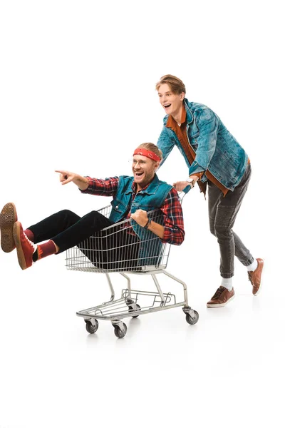 Young hipster man carrying shopping cart with male friend pointing by finger isolated on white — Stock Photo