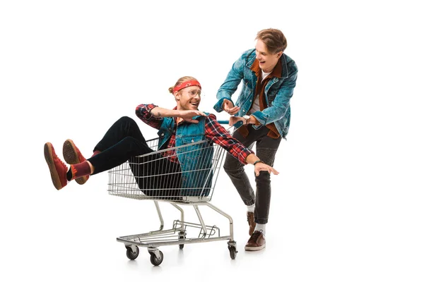 Smiling young man carrying shopping cart with male friend gesturing by hands isolated on white — Stock Photo