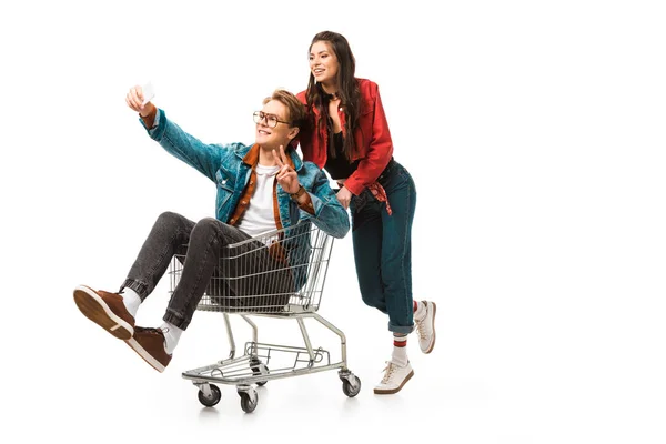 Male hipster in shopping cart doing peace sign and taking selfie while his girlfriend standing behind isolated on white — Stock Photo