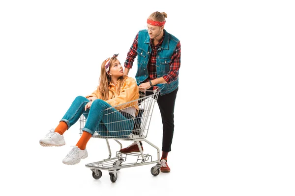 Schöne Hipster-Mädchen sitzt im Einkaufswagen, während ihr Freund hinter isoliert auf weiß stehen — Stockfoto
