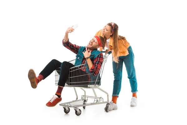 Male hipster in shopping cart doing rock gesture and taking selfie while his girlfriend standing behind isolated on white — Stock Photo