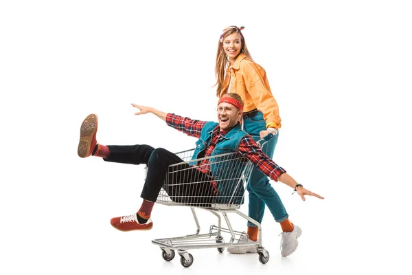 Cheerful hipster girl carrying shopping trolley with happy boyfriend with wide arms isolated on white — Stock Photo