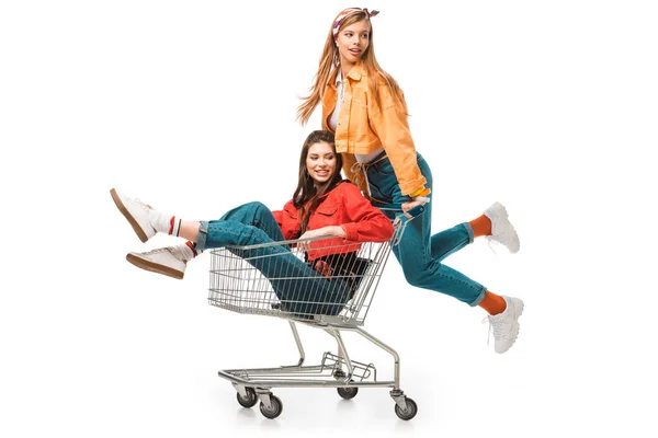 Bela hipster menina carregando carrinho de compras com sorridente amigo feminino isolado no branco — Fotografia de Stock