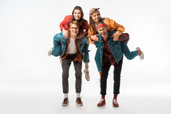 Cheerful stylish hipsters doing piggyback ride to their smiling girlfriends isolated on white — Stock Photo