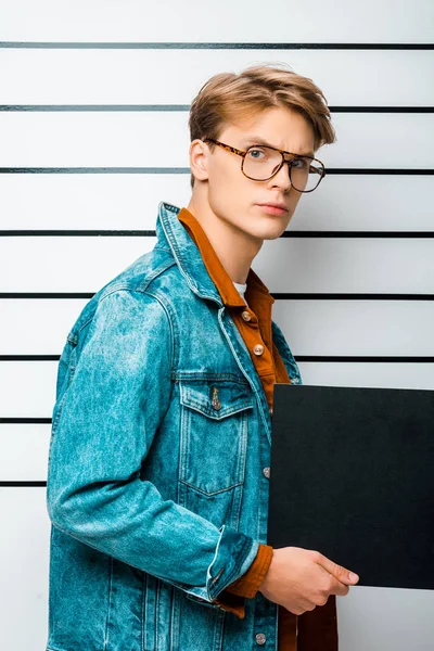 Detenido hipster hombre en gafas con el tablero de la prisión vacío y mirando a la cámara en frente de la línea de policía - foto de stock