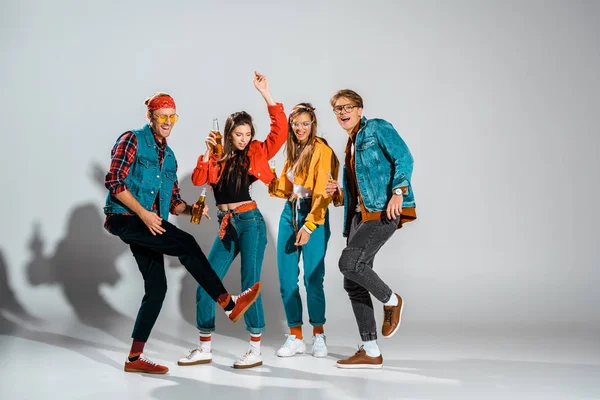 Cheerful stylish hipsters with beer bottles dancing together on grey — Stock Photo