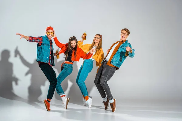 Happy stylish hipsters with beer bottles dancing on grey — Stock Photo
