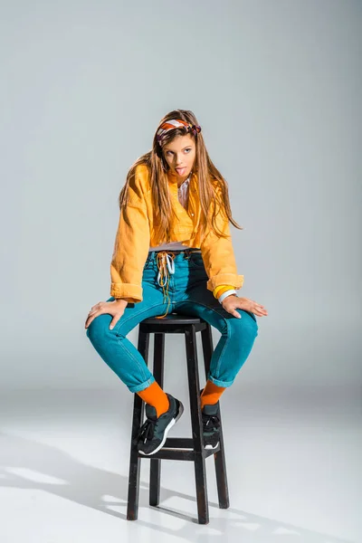 Attractive stylish girl sticking tongue out and sitting on stool on grey — Stock Photo