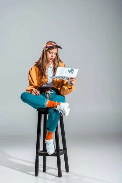 Beautiful girl reading travel newspaper while sitting on stool on grey — Stock Photo