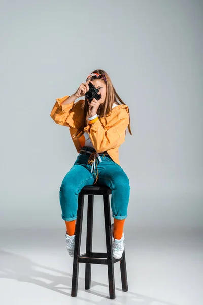 Stylish girl taking photo on camera while sitting on stool on grey — Stock Photo