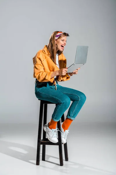 Красива дівчина сміється за допомогою ноутбука і сидить на табуреті на сірому — стокове фото