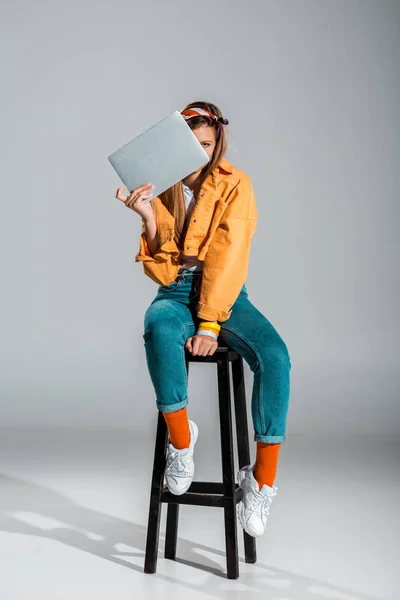 Chica elegante sosteniendo el ordenador portátil mientras está sentado en el taburete en gris - foto de stock