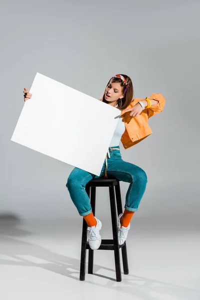Beautiful stylish girl pointing at blank placard on grey — Stock Photo