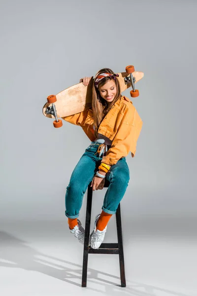 Beautiful smiling girl sitting on stool with skateboard on grey — Stock Photo