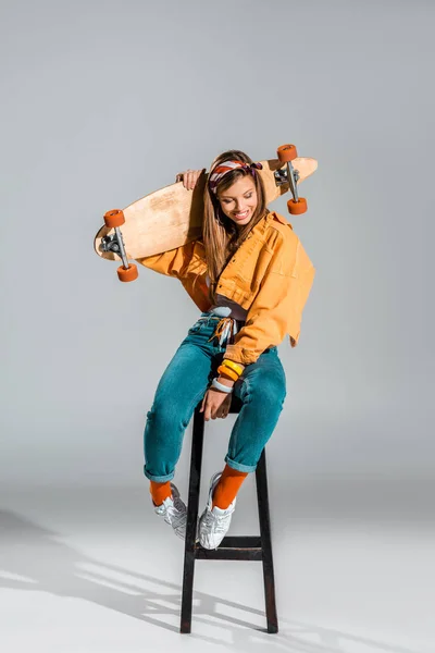 Hermosa chica sonriente sentada en el taburete con monopatín en gris - foto de stock